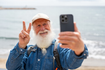cheerful elderly man with a white beard, wearing a beige cap and denim shirt, takes a selfie with his smartphone while making a peace sign near the ocean on a sunny day