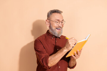 Handsome senior man writing notes in a notebook, appearing happy and content, on a beige background