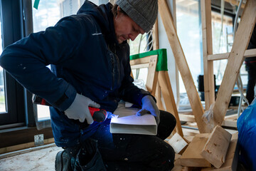 A worker uses a power drill to install a ventilation system inside a house under construction, with...
