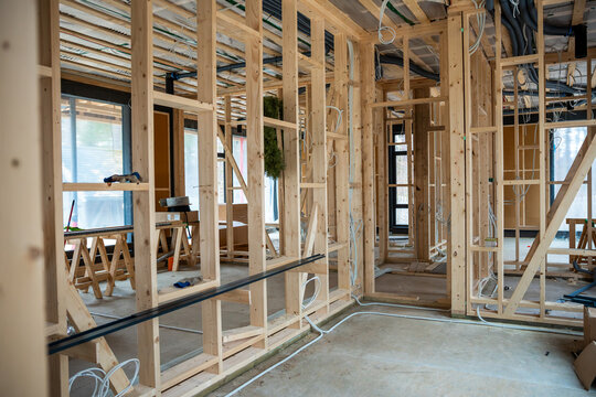 Interior view of a house under construction featuring wooden framing, exposed wiring, ventilation ducts, and natural light through large window openings.