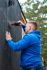 Naklejka premium A man in a blue hoodie operates a power tool on a dark vertical paneled wall of a house under construction, surrounded by tall trees and materials.