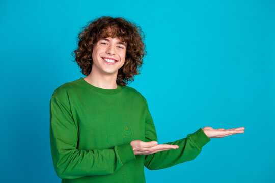 Smiling young man in green sweatshirt presenting with open hands against blue background, confident and welcoming gesture