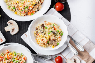Couscous is a dish made from wheat grains (semolina) or millet. Lean couscous with mushrooms, corn, peas, herbs, tomatoes. Side view. Flatlay. Couscous on a light background. Couscous in a white plate