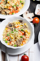 Couscous is a dish made from wheat grains (semolina) or millet. Lean couscous with mushrooms, corn, peas, herbs, tomatoes. Side view. Flatlay. Couscous on a light background. Couscous in a white plate