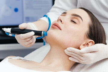 A professional performs a hydropeeling facial treatment on a young woman in a modern salon,...