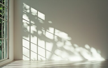 Obraz premium Sunlight streams through a window, casting shadows of leaves and window frames on a plain, light-colored wall above a light wooden floor