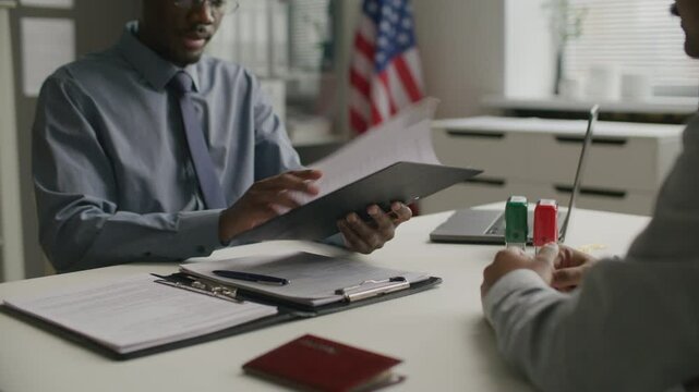African American consular officer taking clipboard from applicant, reviewing documents and asking questions during visa interview at US embassy. Midsection, close-up shot