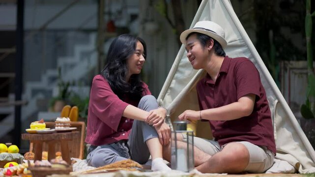 Asian couple sitting close together during a sunny picnic in front of a canvas tent, wearing matching maroon outfits, smiling and having an intimate conversation with desserts and props around