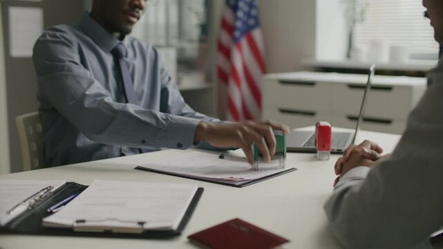Black consular officer stamping approval on visa application form and shaking hands with applicant during meeting at US embassy. Close-up shot