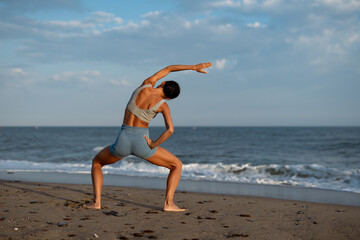 A woman performing a side stretch while holding a wide-legged squat on the beach, showcasing...