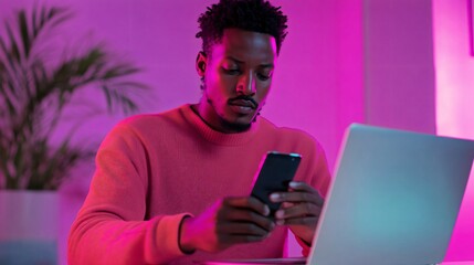 Young man in cozy environment, focused on phone while working on laptop, vibrant lighting and modern decor create an energetic vibe.