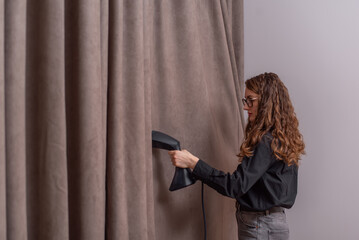 Woman ironing dark beige curtain with hand-held steamer