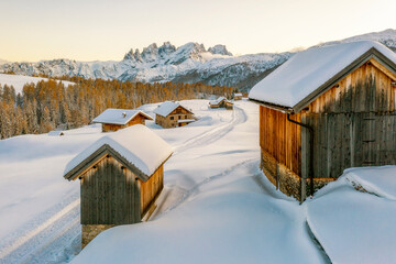 Snowy mountain landscape with wooden cabins in Pian di Fuciade, Trentino, Dolomites. A path leads through the snow to the cabins, with a forest of golden trees and jagged mountains in the background. 