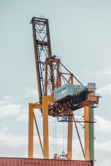 Large industrial port crane lifting a shipping container against a bright sky. The towering yellow...