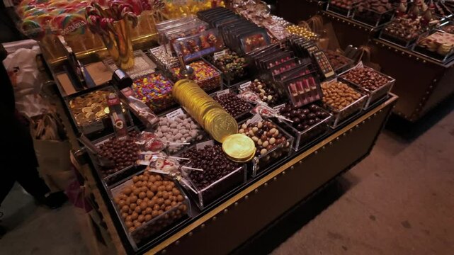 A wide shot of a chocolate and candy stall at Mercat de la Boqueria, Barcelona, showcasing golden foil-wrapped chocolate coins, chocolate nuts, lollipops, and novelty sweets in warm lighting.