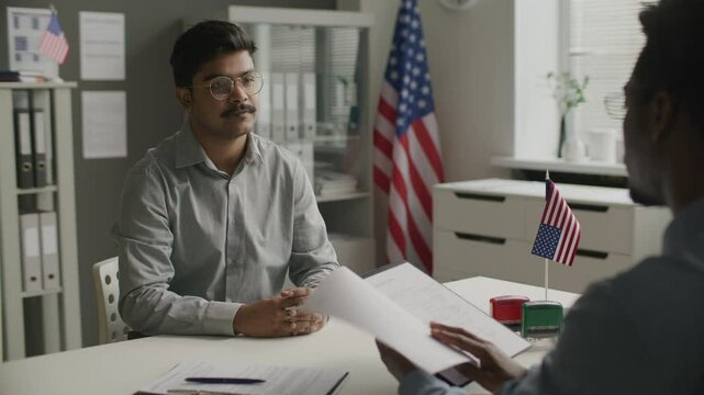 Indian man sitting at desk with American flag on it, answering questions during visa interview while immigration officer checking his documents at US Embassy