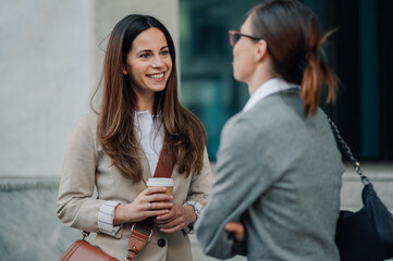 Two businesswomen talking and smiling outdoors while holding coffee