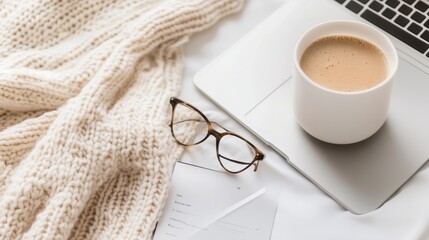 Cozy workspace featuring a cup of coffee, laptop, glasses, and a warm knitted blanket on a soft surface at home