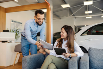 Friendly smiling car dealer standing at car showroom and giving a contract to a female buyer.