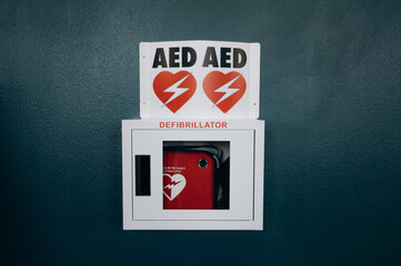 In a gym, a defibrillator is mounted on a dark wall in a protective box. A bright red sign on the device attracts attention, reminding you of the importance of safety during intense workouts.
