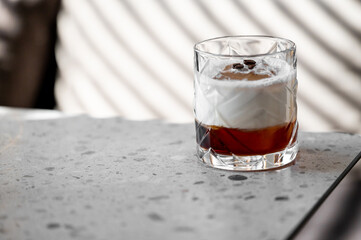 A glass of iced coffee topped with cream, placed on a gray speckled surface with a blurred striped background, garnished with coffee beans for a refreshing, elegant look.