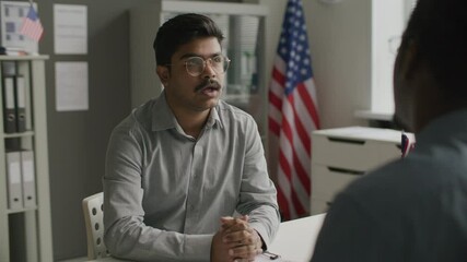 Indian man sitting at office desk, talking to consul during visa interview in embassy with American flag in background. Over the shoulder view