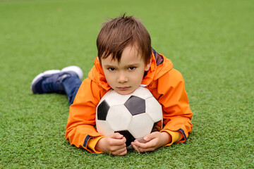 Obraz premium Young boy in orange jacket lies on football field, hugging soccer ball with serious or sad look. Maybe his team lost. Passion for sports, childhood dreams, and competition emotions concept 