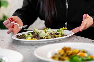 A white plate with a fresh salad of cucumber slices, mixed greens, and yellow flowers. A hand with red-painted nails holds a fork, set on a light table.