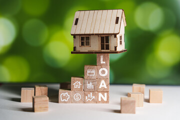 A small wooden house model is precariously balanced on a stack of wooden blocks spelling LOAN....