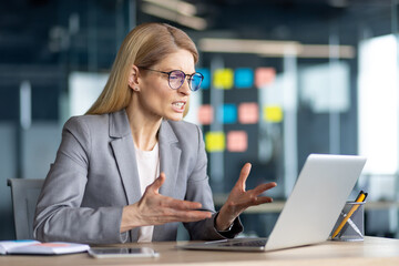 A businesswoman is sitting at her desk and having a heated discussion online using her laptop in a...