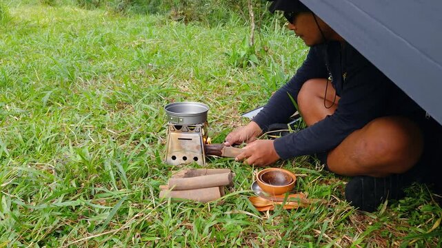 A male adventurer is cooking sauce to make a burger under a flysheet in the dense forest. The concept of bushcraft camping in a dense pine forest for adventure and cooking with simple equipment