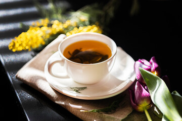 White cup with green tea on a dark background in sunlight. White porcelain cup with saucer. Yellow flowers nearby. Large green tea leaves unfolded in the cup.