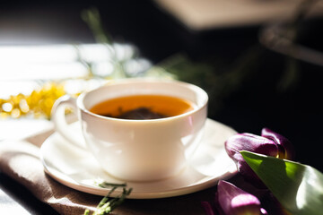 White cup with green tea on a dark background in sunlight. White porcelain cup with saucer. Yellow flowers nearby. Large green tea leaves unfolded in the cup.