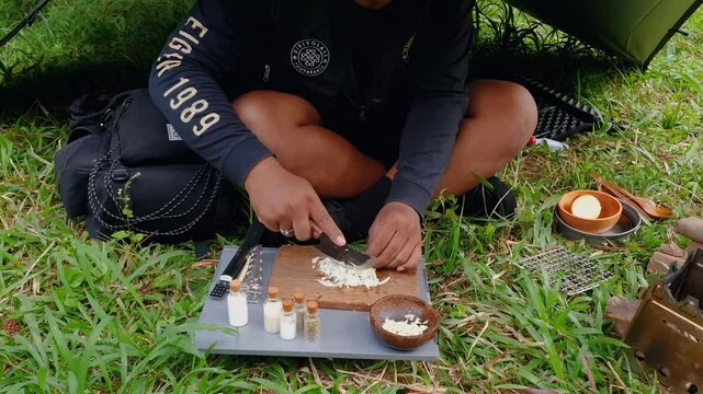 A male adventurer is cooking sauce to make a burger under a flysheet in the dense forest. The concept of bushcraft camping in a dense pine forest for adventure and cooking with simple equipment