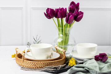 Still life of white porcelain cups and teapot, tulips in a glass vase, mimosa flowers, light background. Tea party. Spring tea in good weather.