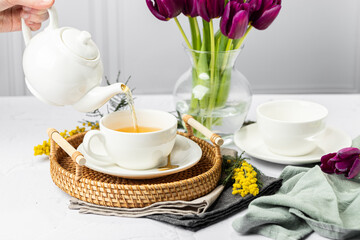 Green tea is poured from a white teapot into white porcelain cups. In the background are tulips in...