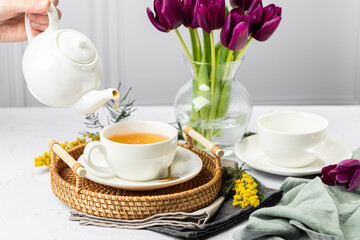 Green tea is poured from a white teapot into white porcelain cups. In the background are tulips in a glass vase, mimosa flowers, light background. Tea drinking.