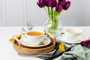 Green tea in a white porcelain cups and teapot, tulips in a glass vase, mimosa flowers, light background. Tea party.