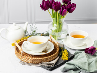 Green tea in a white porcelain cups and teapot, tulips in a glass vase, mimosa flowers, light background. Tea party.