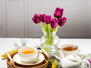 Green tea in a white porcelain cups and teapot, tulips in a glass vase, mimosa flowers, light background. Tea party.