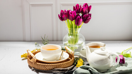 Green tea in a white porcelain cups and teapot, tulips in a glass vase, mimosa flowers, light background. Tea party.