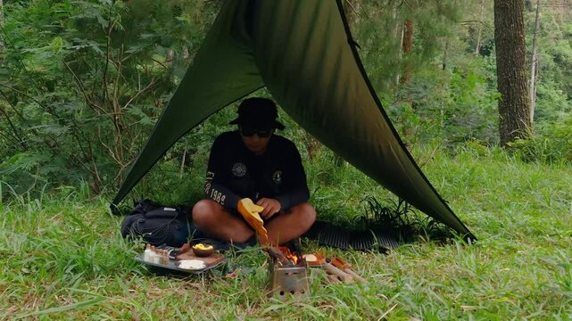 A male adventurer is cooking sauce to make a burger under a flysheet in the dense forest. The concept of bushcraft camping in a dense pine forest for adventure and cooking with simple equipment