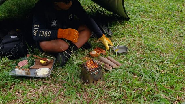 A male adventurer is cooking sauce to make a burger under a flysheet in the dense forest. The concept of bushcraft camping in a dense pine forest for adventure and cooking with simple equipment