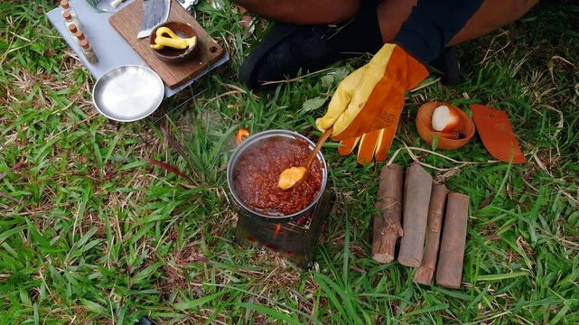 A male adventurer is cooking sauce to make a burger under a flysheet in the dense forest. The concept of bushcraft camping in a dense pine forest for adventure and cooking with simple equipment