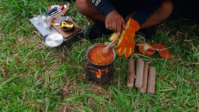 A male adventurer is cooking sauce to make a burger under a flysheet in the dense forest. The concept of bushcraft camping in a dense pine forest for adventure and cooking with simple equipment