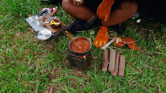A male adventurer is cooking sauce to make a burger under a flysheet in the dense forest. The concept of bushcraft camping in a dense pine forest for adventure and cooking with simple equipment