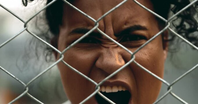 One upset young black woman yelling behind metal fence barrier looking at camera grimacing in anger and screaming in despair. 20s person feeling outrage