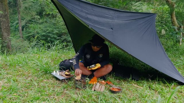 A male adventurer is cooking sauce to make a burger under a flysheet in the dense forest. The concept of bushcraft camping in a dense pine forest for adventure and cooking with simple equipment