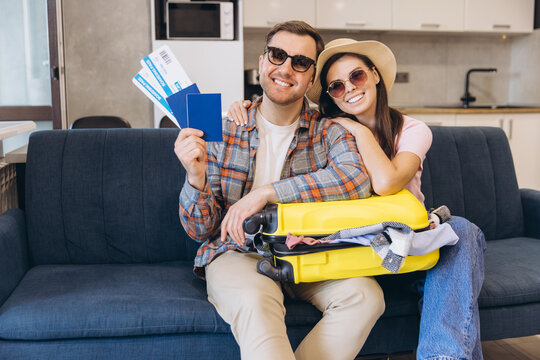 Happy couple showing passport and tickets sitting on sofa with suitcase ready for vacation