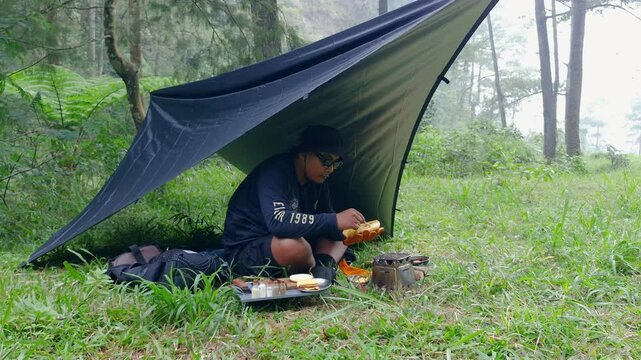A male adventurer is cooking sauce to make a burger under a flysheet in the dense forest. The concept of bushcraft camping in a dense pine forest for adventure and cooking with simple equipment